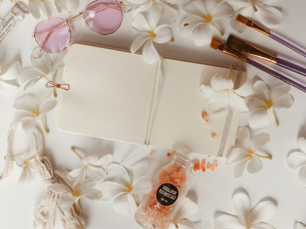 Flat lay featuring an open journal, plumeria flowers, paint brushes, and Himalayan rock salt for creative inspiration.