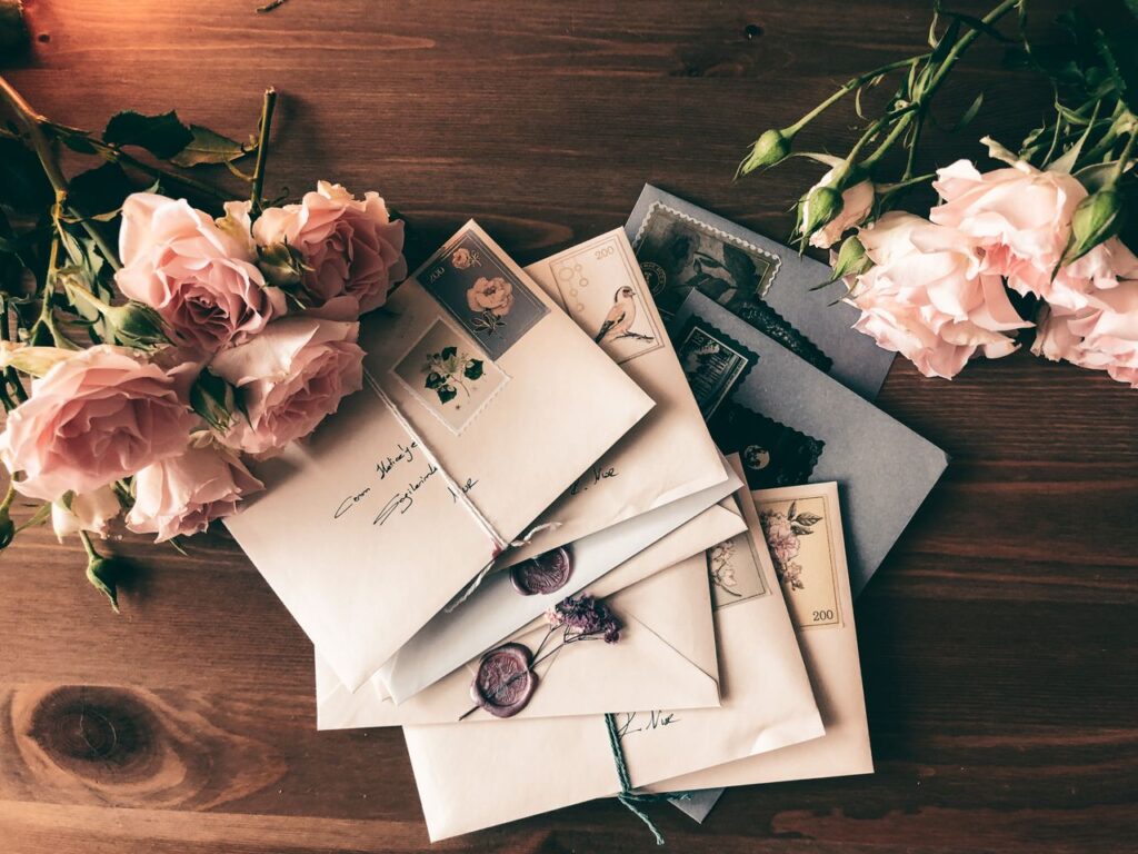 Top view composition of stack of letters and pink roses placed on wooden table in daytime